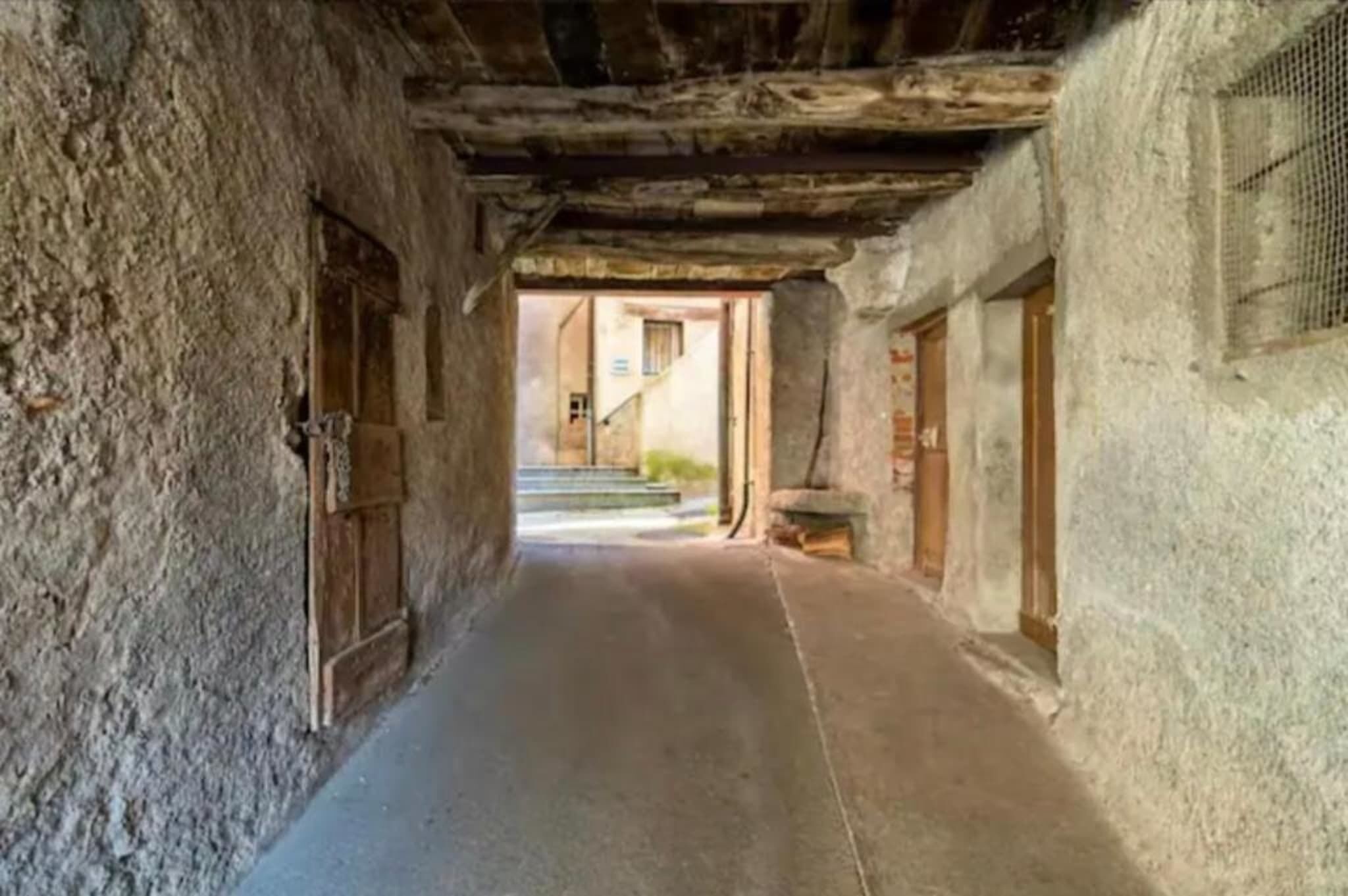 Un estrecho pasillo con paredes de piedra tosca, puertas de madera y una escalera que sube a un nivel superior.