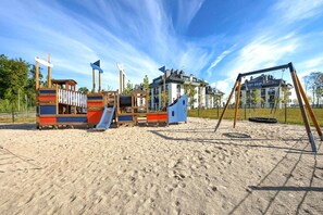 Ein Spielplatz mit verschiedenen Spielgeräten, darunter Rutschen und Kletterbereichen, umgeben von einem Sandbereich und Wohnhäusern im Hintergrund.