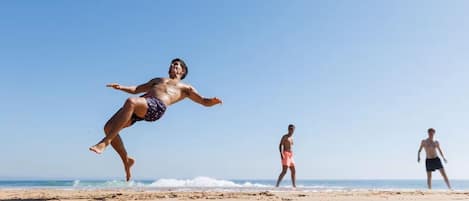 Une scène de plage où l’on voit une personne sauter pour attraper un ballon, une autre qui marche et une troisième qui se tient à l’arrière-plan.