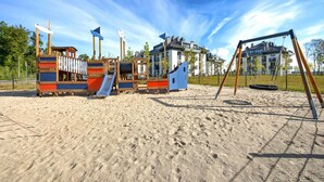 En lekplats med olika typer av lekanord, inklusive rutschkanor och klätterställningar, omgiven av ett sandområde och bostadshus i bakgrunden.