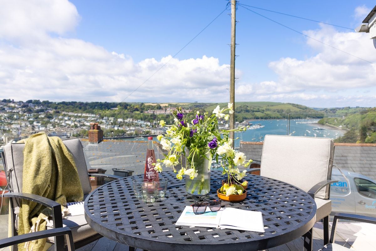 A round table with a bottle, a vase with flowers, and a bowl on a balcony overlooking a town and water.