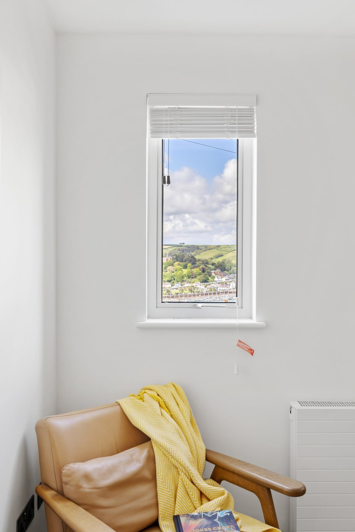 A room with a window overlooking a landscape, a chair with a yellow blanket, and a white radiator.