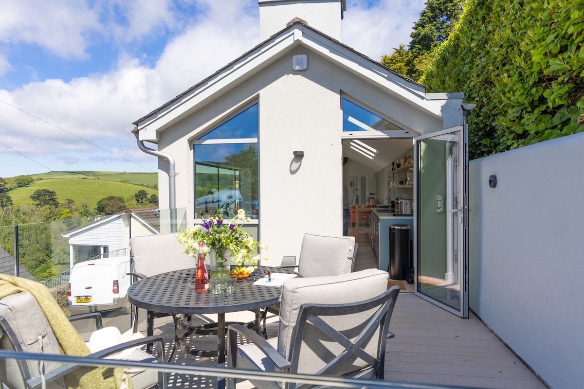 A modern house with a balcony featuring a dining table and chairs, a glass door leading to a kitchen, and a view of the countryside.