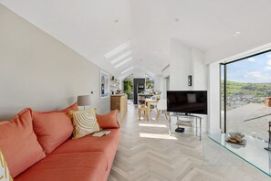 A modern living room with a sofa, a glass coffee table, and a TV. There is a view of a cityscape through large windows.