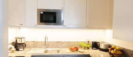 Una cocina moderna con microondas integrado, electrodomésticos de acero inoxidable, un lavabo y una encimera con frutas y una cafetera.
