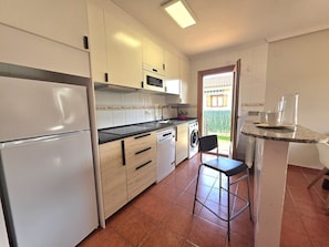 Cocina compacta con refrigerador, horno, microondas, lavavajillas y un pequeño comedor con una silla.