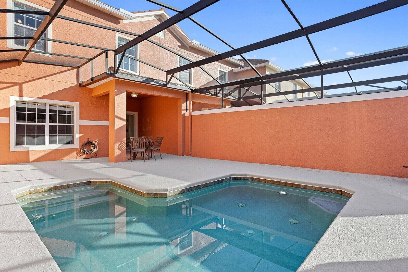 A swimming pool with a clear view of the sky, a covered patio area with a table and chairs, and a building with orange walls and a window.