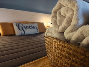 A bed with a dark brown mattress, a woven basket with a rolled-up white towel, and a pillow with 'Gracias' written on it. - Hotel Alta Mendoza (Mendoza)