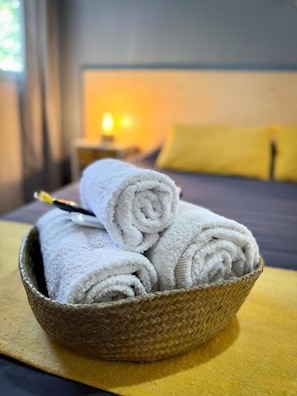 A bed with a yellow pillow and a headboard, a bedside table with a lamp, and a woven basket containing rolled white towels. - Hotel Alta Mendoza (Mendoza)