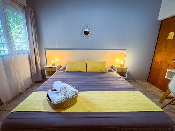 Hotellihuone, jossa on sänky, kaksi yöpöytää valaisimineen, sängynpääty, ikkuna verhoineen ja puuovi.