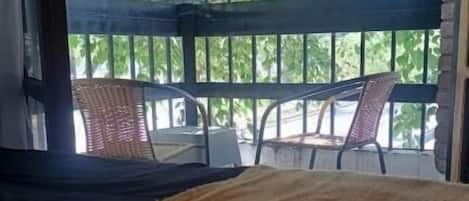 A room with a bed, a wicker hanging chair, and a view of greenery through a window with metal bars.