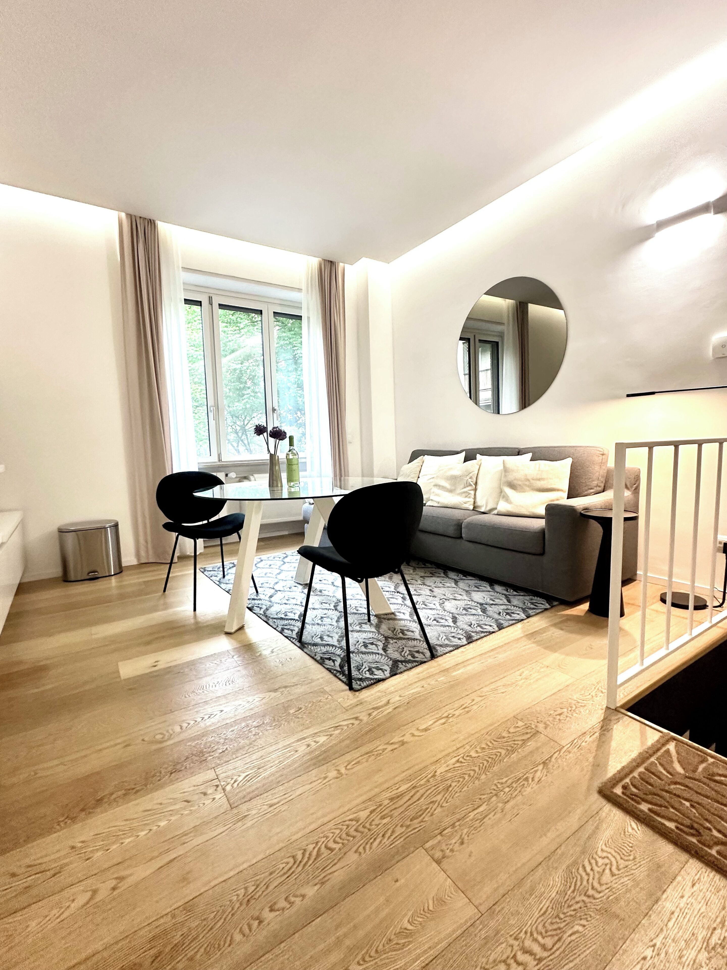 Ein modernes Wohnzimmer mit einer Couch, einem Esstisch mit Stühlen, einem großen Fenster und einem Spiegel an der Wand.