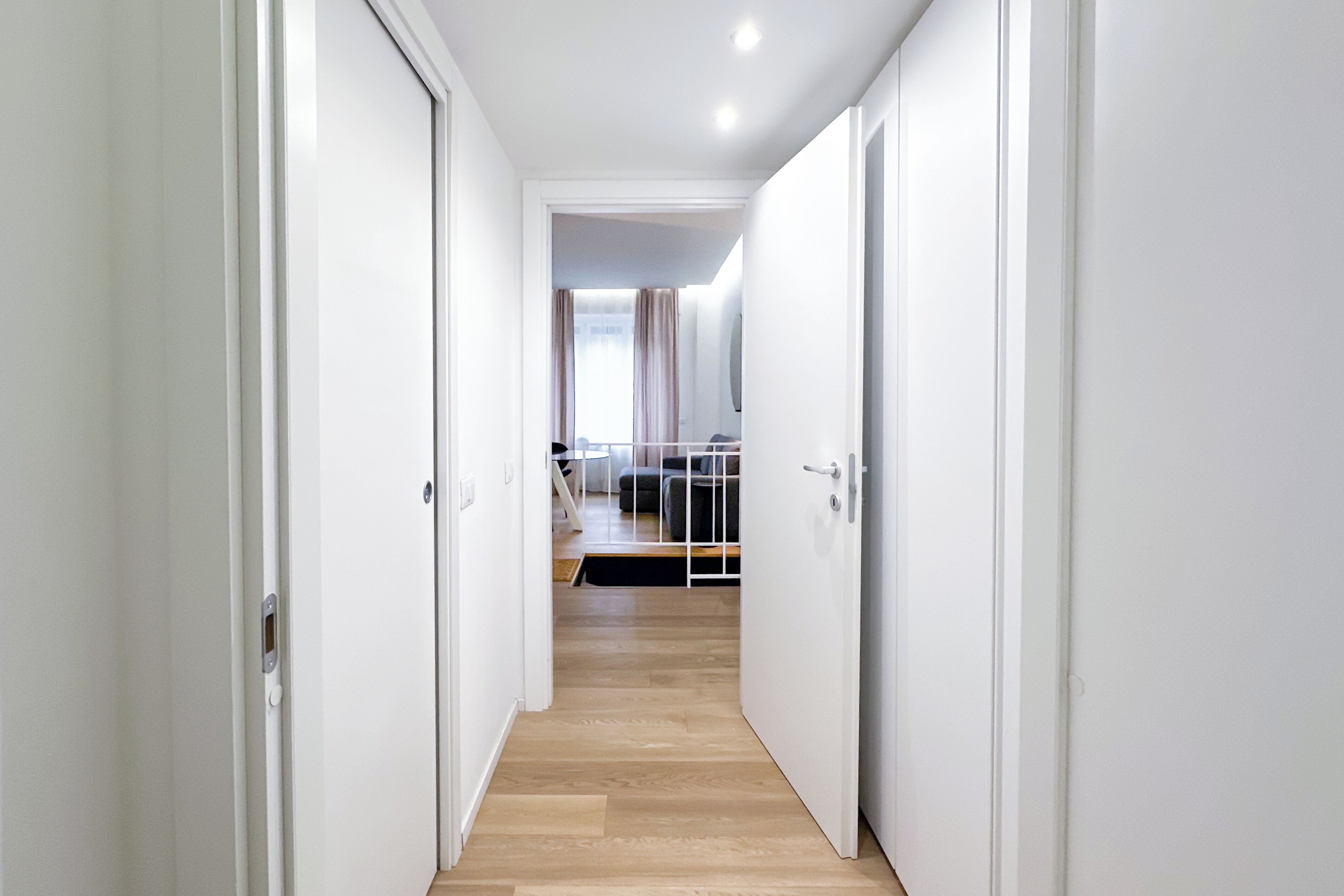 A narrow hallway with white doors leading to a bedroom with a bed and a window.