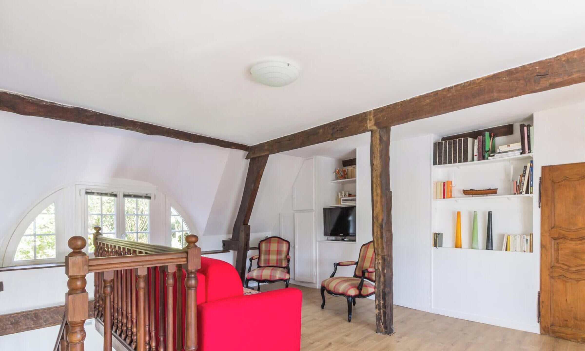 Een kamer met een houten trap, een rode bank, een geruite fauteuil, een boekenkast en een deur.