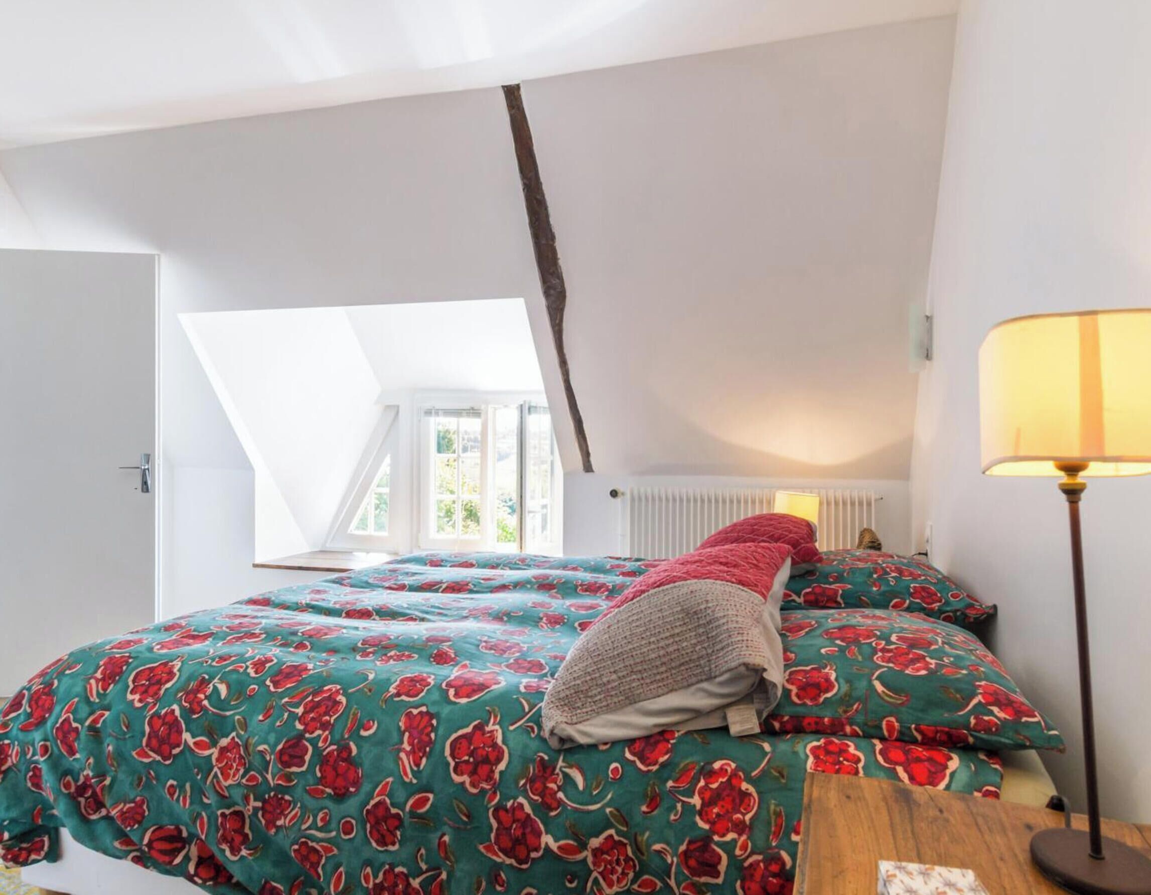 Un dormitorio con techo inclinado, una cama con ropa de cama floral, una lámpara de noche y vistas a través de una ventana abierta.