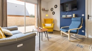Un salon moderne comprenant un canapé, un fauteuil jaune, une chaise bleue, un téléviseur et une porte-fenêtre coulissante donnant sur une terrasse.