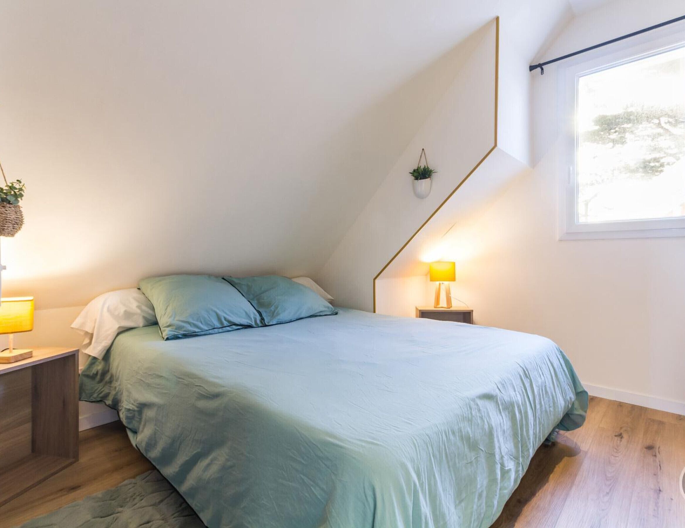 Een slaapkamer met een bed, twee nachtkastjes, een hangplant en een raam.