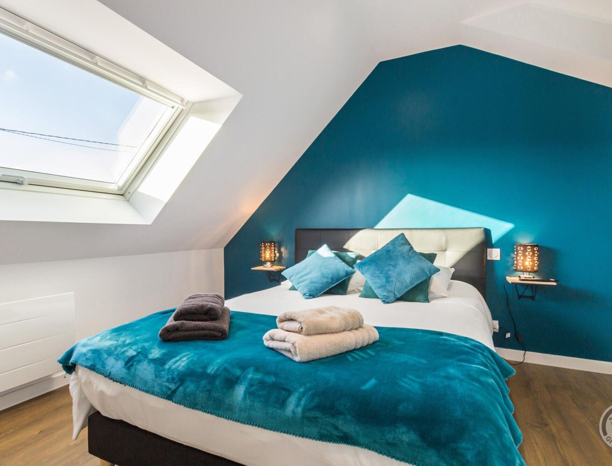 A bedroom with a sloped ceiling, a skylight, a bed with a teal bedspread, two bedside lamps, and a nightstand.