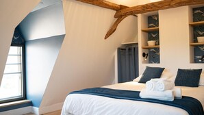 A bedroom with a sloped ceiling, a bed with white bedding and dark pillows, a wooden beam structure, and a wall with shelves holding decorative items.