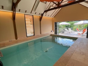Une piscine intérieure donnant sur une terrasse extérieure aménagée avec des chaises longues et des parasols.