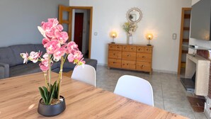 Un coin repas avec une table en bois, des chaises, une orchidée en pot, un meuble en bois sur lequel sont fixées deux lampes murales, et un téléviseur posé sur un meuble.