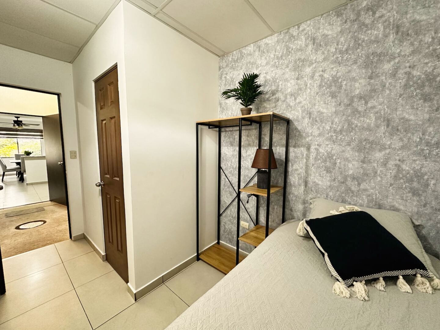 A bedroom with a bed, a small wooden shelf with a lamp, a potted plant, and a door leading to a bathroom.