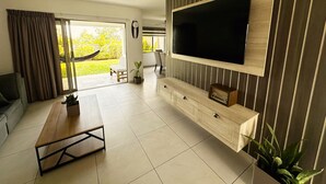 Ett modernt vardagsrum med en stor TV, ett soffbord i trä, en soffa och en skjutdörr i glas som leder ut till en trädgård.