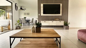 Ett modernt vardagsrum med ett soffbord i trä, en TV som är monterad på en väggpanel i trä, en matplats med stolar och ett bord samt en soffa.