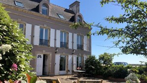 A two-story building with a balcony, a door, and a garden area with plants and outdoor furniture. - Detached house with panoramic view - 8 guests (Portbail)