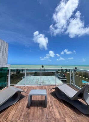 Ein Balkon mit zwei Liegestühlen und einem kleinen Tisch, mit Blick auf einen Strand und das Meer.
