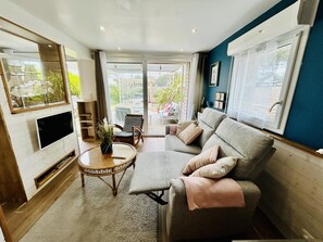 Een moderne woonkamer met een grijze bank, een flatscreen televisie aan de muur, een rieten salontafel en een grote schuifdeur naar een buitenruimte.