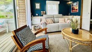 Un salón acogedor con un sofá gris, una silla de madera, una mesa redonda de mimbre y un ventanal grande.