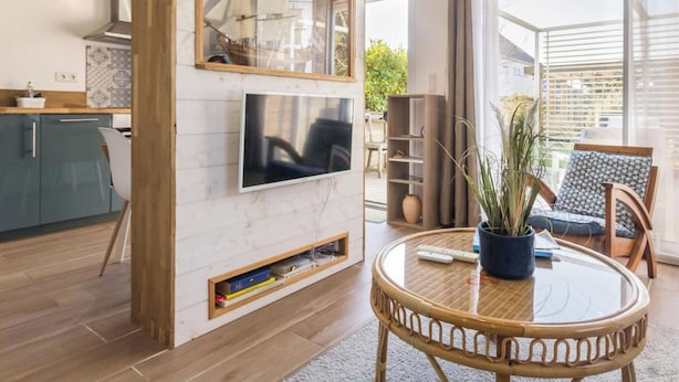 Una sala moderna con un televisor montado en una pared blanca, una mesa redonda de mimbre y una zona de estar con una silla estampada.