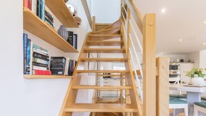 Eine Holztreppe mit Geländer, die zu einem Küchenbereich mit weißen Schränken und einem Esstisch führt.