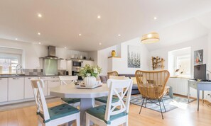 A modern kitchen with white cabinets, a dining table with chairs, a large window, and a hanging light fixture. - Charming duplex in Portbail - 2 guests (Portbail)