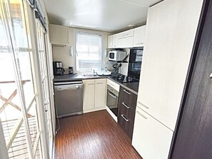 A compact kitchen with a dishwasher, oven, microwave, and a window with blinds. - Family mobile home with AC - 4 guests (Bourg-Charente)