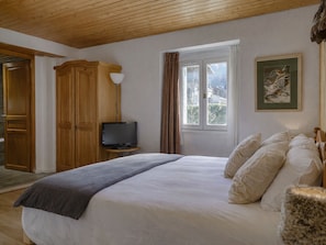 Ein Schlafzimmer mit einem Bett, einem Fernseher auf einem kleinen Tisch, einem Fenster mit Vorhängen und einer Holzbalkendecke.
