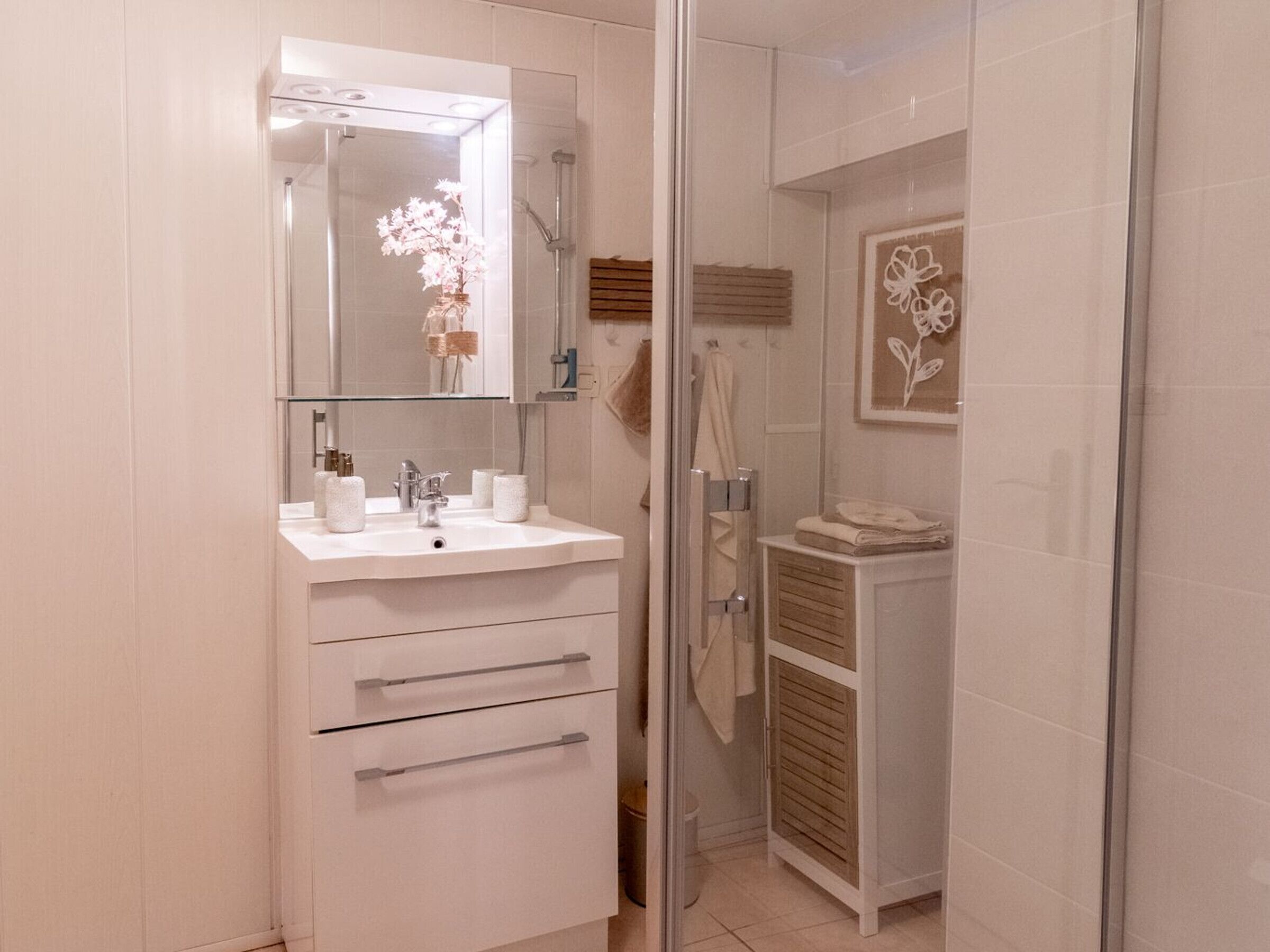 Une salle de bain avec un meuble-lavabo blanc, un miroir avec éclairage intégré, une douche et une œuvre d’art florale encadrée accrochée au mur.