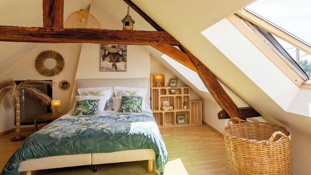 Un dormitorio con techo inclinado, vigas de madera, cama con colcha de temática tropical, una cesta de mimbre y un tragaluz.