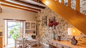 Un interior rústico con paredes de piedra vista, vigas de madera, una zona de comedor con muebles blancos y una escalera.