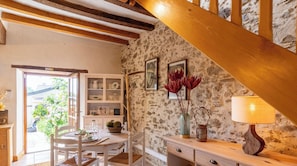 Un intérieur rustique avec des murs en pierre apparentes, des poutres en bois, un coin repas avec des meubles blancs et un escalier.