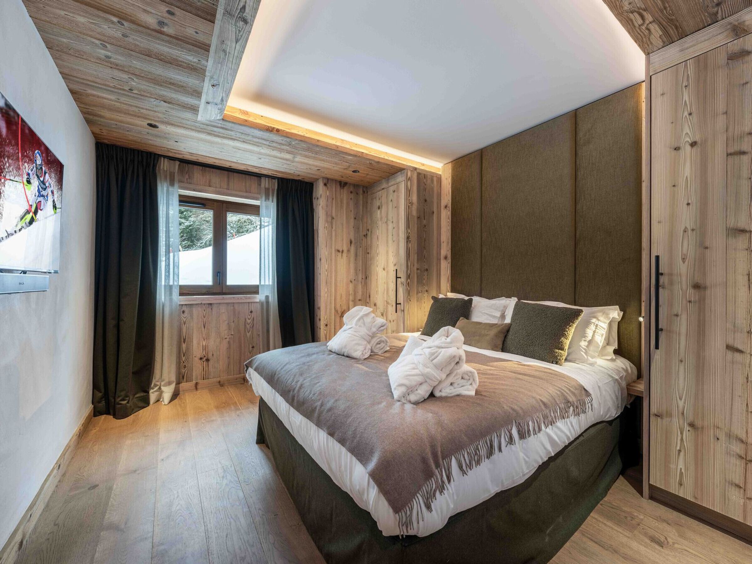 A bedroom with a wooden ceiling, a large bed with pillows, a window with curtains, and a painting on the wall.