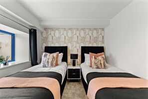 A hotel room with two beds, a nightstand with a lamp, a window with a view, and a patterned wall. - Cozy Leeds Hideaway Near the City (Leeds)