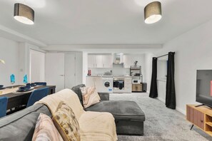 Ein modernes Wohnzimmer mit einem grauen Ecksofa, einem Essbereich mit blauen Stühlen, einer Küche mit weißen Schränken und einem Fernseher auf einem Holzboden.