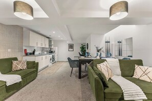 Ein modernes Wohnzimmer mit grünen Sofas, einem Esstisch und einer Küche im Hintergrund.