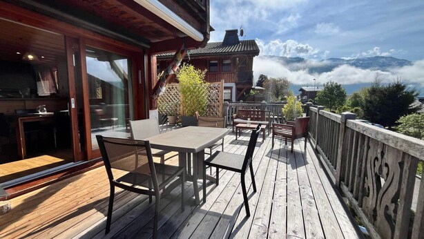 A wooden deck with outdoor seating, a glass door entrance, and a view of mountains. - Apartment with panoramic view - 4 guests (Samoëns)