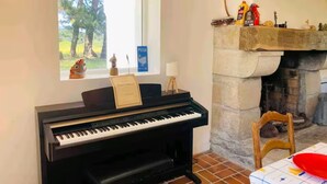 Ett piano, ett fönster med utsikt över träd, en öppen spis och ett litet bord med ett rött föremål på.