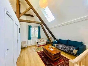 Ein Loft-Zimmer mit schrägem Dach, einer Couch, einem Couchtisch, einem Sessel und einem fenster mit Vorhängen.