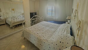 Ein Schlafzimmer mit Bett, Fernseher, Fenster mit Vorhängen und einem Spiegel.