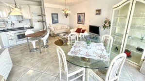 Eine moderne Küche mit weißen Schränken, ein Essbereich mit einem Glastisch und ein Wohnzimmer mit einer Couch und Fernseher.